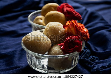 Picture of delicious Rawa laddu decorated in bowl with fresh red roses in a blue background. Rawa laddus made during diwali festival in India.