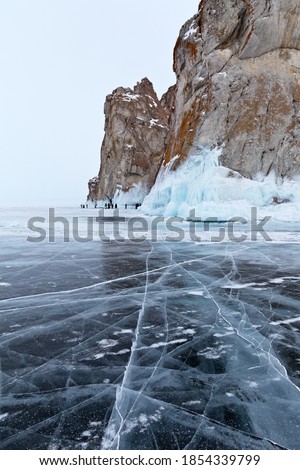 Beautiful winter landscape of frozen Lake Baikal on cold February day. Blue transparent ice with cracks near Rocks of Olkhon Island. A group of tourists take pictures of the icy rocks. Winter travel