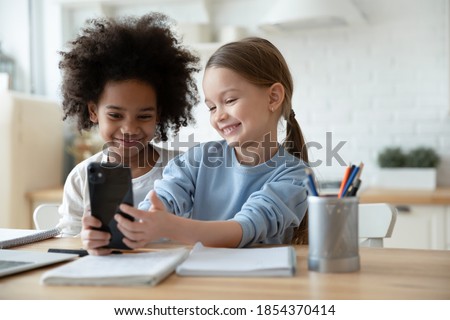 Two happy diverse little girls sisters having fun with smartphone together, distracted from studying, smiling cute kids looking at phone screen, taking selfie, watching cartoons, multiracial family