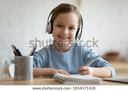 Head shot portrait smiling little girl wearing headphones looking at camera, sitting at table, studying at home, happy pretty child doing homework, holding pen, writing notes, watching webinar