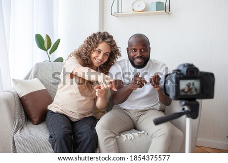 Nice multinational couple shoots a professional video for their blog. Curly expectant mother and African-American husband joyfully gesticulate. Pregnancy, family and technology concept.