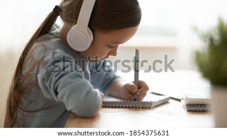 Close up side view little girl wearing headphones writing in notebook, cute child schoolgirl doing homework, making notes during online lesson, sitting at table at home, homeschooling concept