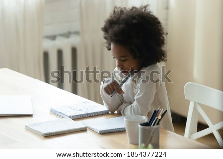 Thoughtful focused African American little girl studying at home, sitting at table, holding pen, pensive dreamy girl child pondering difficult school tasks, doing homework, homeschooling concept Royalty-Free Stock Photo #1854377224