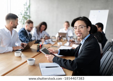 Corporate Meeting. Happy Asian Businessman Using Digital Tablet Smiling Looking At Camera Sitting With Colleagues At One Desk In Modern Office. Free Space For Text, Selective Focus