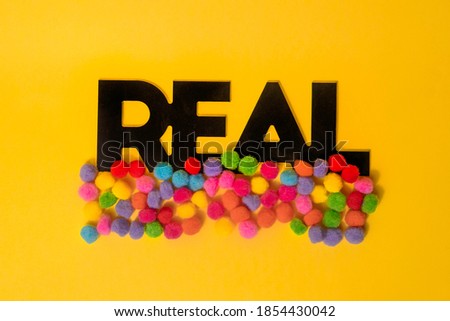 Word ''Real'' covered with colorful pom poms on a yellow background.