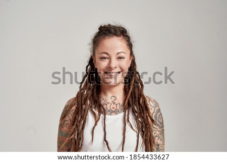 Portrait of young tattooed caucasian woman with dreadlocks wearing white shirt, smiling at camera while posing isolated over grey background. Front view. Horizontal shot Royalty-Free Stock Photo #1854433627