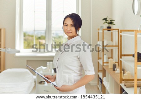 Portrait of serious Asian cosmetologist or aesthetic nurse in her workplace. Professional beautician with clipboard standing in beauty salon. Female massagist in massage room looking at camera Royalty-Free Stock Photo #1854435121
