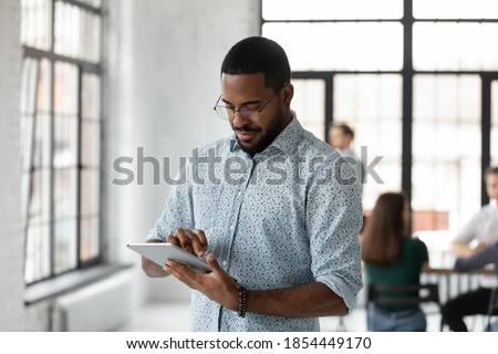 Easy communication. Busy concentrated young black male employee manager corporate worker standing at office open space focused on tablet pc screen accepting urgent order reading email texting answer Royalty-Free Stock Photo #1854449170