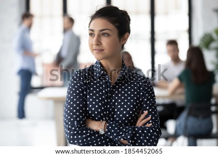 Professional look. Confident young female of indian ethnicity capable specialist standing in office space with arms crossed on chest looking at distance taking rest of routine work thinking pondering