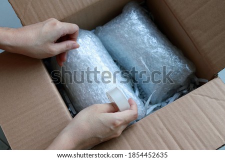 Woman’s hands packing fragile items with plastic air bubble wrapped and adhesive tape and putting in a brown cardboard parcel box. Royalty-Free Stock Photo #1854452635