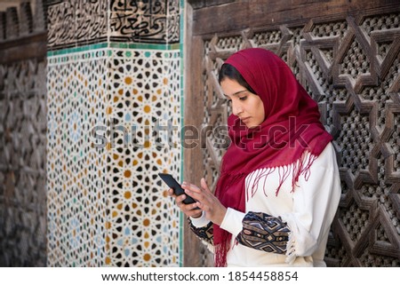 Muslim woman writing a message on her smart phone in traditional clothing with red headscarf on her head beside a traditional arabesque Moroccan wall