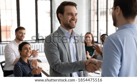 Happy motivated millennial man intern is being hired on regular job employed to international company, loyal young male manager handshaking with leader employer on formal meeting with corporate team Royalty-Free Stock Photo #1854475516