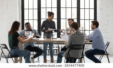 Training handouts. Qualified biracial female mentor trainer giving millennial trainees students paper documents by subject matter to educate, indian woman team leader setting problems to diverse staff Royalty-Free Stock Photo #1854477400