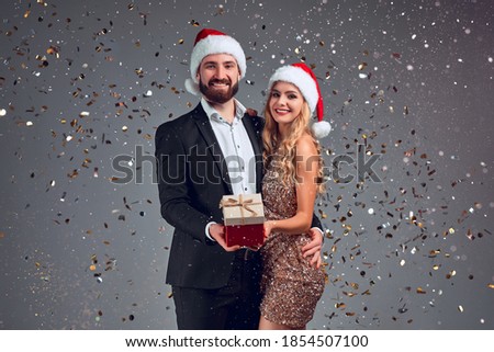 Merry Christmas. Cheerful couple holding Xmas gift box smiling to camera standing over grey background, wearing red Santa hats. New Year presents. Winter holidays celebration.