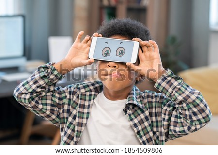 Cute little mixed-race boy in white t-shirt and checkered shirt holding smartphone with funny eyes on screen in front of his face