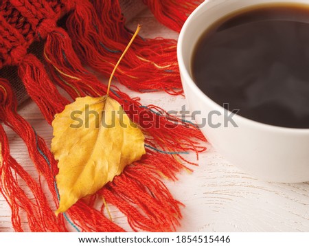 Close-up of dry yellow fall leaf on a knitted red fringed scarf near white mug of hot steaming coffee. Cozy autumn mood concept. Selective focus. Top view. 