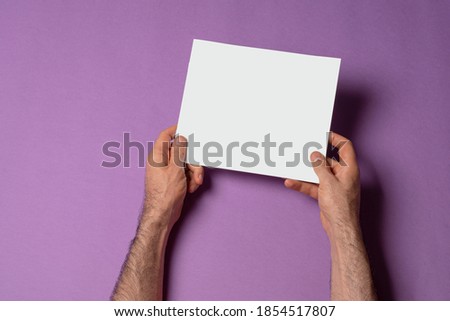 Male hands holding a square proportion catalog with blank cover on purple background, mock-up series template ready for your design, cover selection path included.