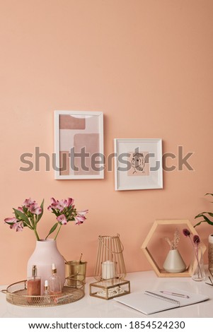 Workplace of interior designer by pink wall with two pictures in frames by table with flowers in vases, beauty care products, candles and other stuff