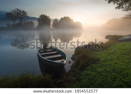 Calm scenic picture of small fishing boat on the Adda river on a hazy sunrise with foliage