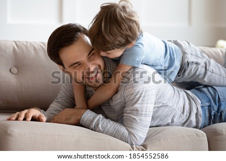 Close up overjoyed loving father and adorable son hugging, cuddling, relaxing on couch together, cute little boy lying on dad back, happy family enjoying tender moment, leisure time on weekend