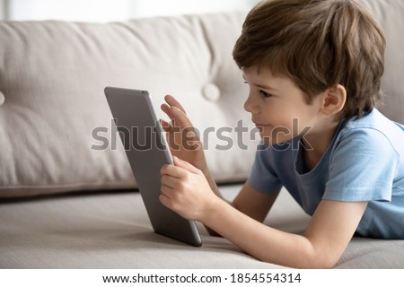 Close up little boy using computer tablet, lying on cozy couch at home alone, cute kid looking at electronic device screen, watching cartoons, playing game, browsing gadget apps, chatting online