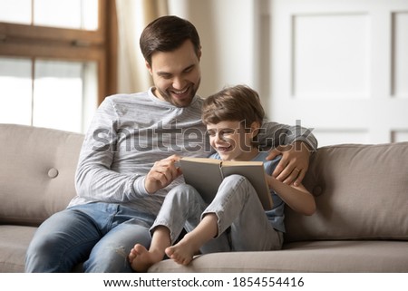 Loving happy father teaching adorable son to read, smiling dad and little boy child hugging, sitting on couch, holding book with fairy tale story, family spending weekend together, leisure time Royalty-Free Stock Photo #1854554416