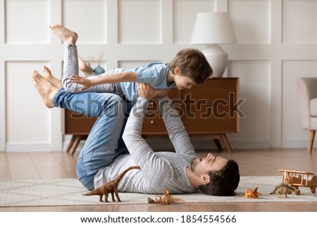 Overjoyed loving father playing active game with adorable son, lying on warm floor at home, happy dad holding carrying little boy with hands outstretched pretending flying, enjoying leisure time Royalty-Free Stock Photo #1854554566