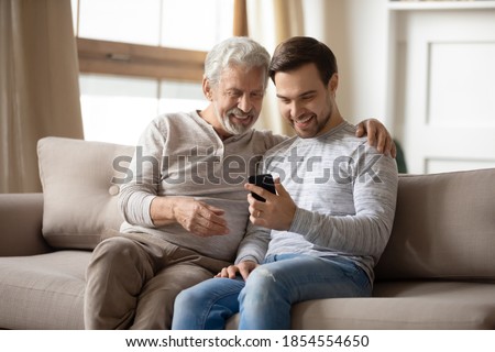 Happy older father and adult son hugging, using phone at home together, smiling mature grandfather and grandson looking at screen, browsing apps, young man teaching senior dad to use smartphone Royalty-Free Stock Photo #1854554650