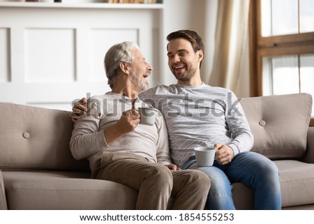 Overjoyed mature father and adult son drinking tea together, chatting, enjoying pleasant conversation, happy older grandfather and grandson laughing at funny joke, sitting on couch at home Royalty-Free Stock Photo #1854555253