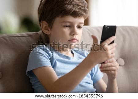 Close up cute little boy using smartphone, looking at screen, curious child holding phone in hands, sitting on couch at home alone, playing mobile device game, watching cartoons online, chatting