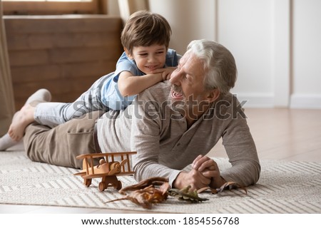 Overjoyed mature grandfather playing with adorable grandson at home, older man lying on warm floor with cute little boy grandchild on back, family spending leisure time together, having fun Royalty-Free Stock Photo #1854556768