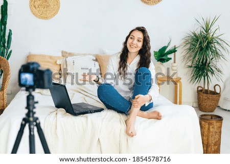 The blogger girl sits on the bed in the bedroom and records a video clip on the camera.