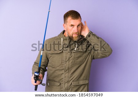 Senior fisherman isolated on purple background trying to listening a gossip.