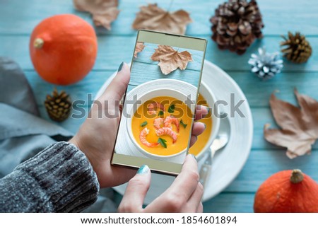 Woman taking snapshot with her smartphone of shrimp and pumpkin soup on blue table