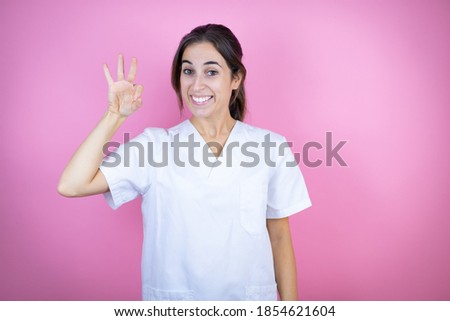 Young brunette doctor girl wearing nurse or surgeon uniform over isolated pink background doing ok sign with fingers and smiling, excellent symbol