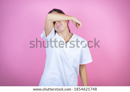 Young brunette doctor girl wearing nurse or surgeon uniform over isolated pink background covering eyes with arm smiling cheerful and funny. Blind concept.