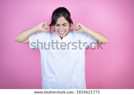 Young brunette doctor girl wearing nurse or surgeon uniform over isolated pink background covering ears with fingers with annoyed expression for the noise of loud music. Deaf concept.