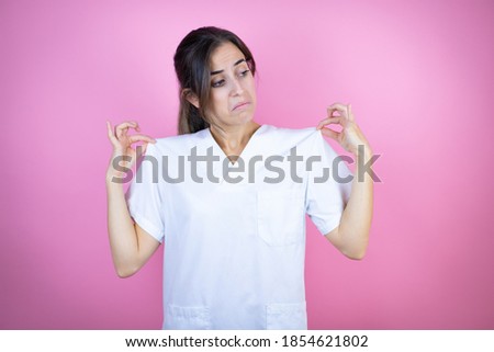 Young brunette doctor girl wearing nurse or surgeon uniform over isolated pink background holding her t-shirt with a successful expression