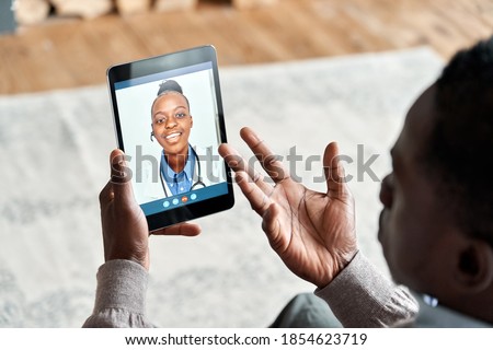 Over shoulder view of african man video calling female virtual doctor consulting patient on tablet at home. Online telemedicine chat visit meeting. Ehealth, telehealth consultation, tele medicine. Royalty-Free Stock Photo #1854623719