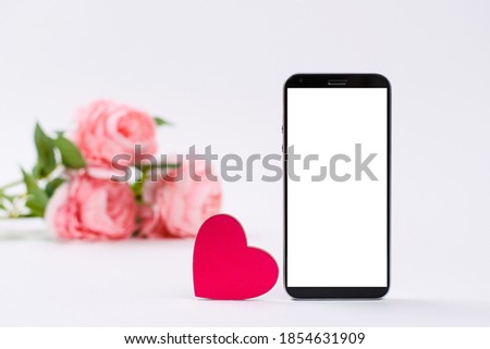 Pink heart and smartphone with blank white display against white background. in the background a bouquet of pink roses. Concept of declaration of love through messages