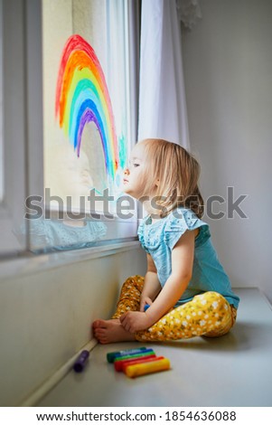 Adorable toddler girl drawing rainbow on window glass as sign of hope. Creative games for kids staying at home during lockdown. Self isolation and coronavirus quarantine concept