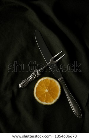 A closeup vertical shot of a silver fruit fork and knife with  a fresh orange slice on a dark silk background