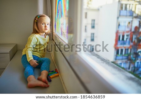Adorable toddler girl drawing rainbow on window glass as sign of hope. Creative games for kids staying at home during lockdown. Self isolation and coronavirus quarantine concept