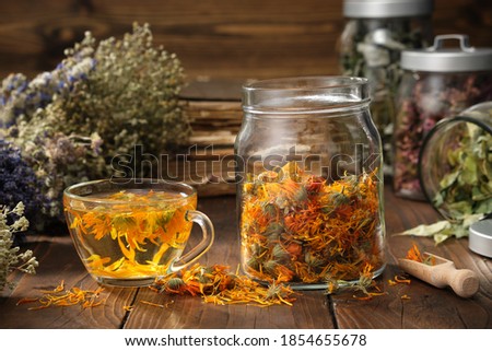 Cup of healthy marigold tea, glass jar of dry calendula flowers. Jars of medicinal herbs and old books on background.  Alternative medicine.