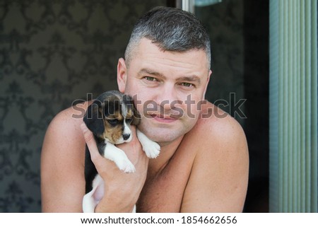 close-up portrait of happy senior man looking at camera on a home background of window. Man holding and hugging beagle dog. Smiling, enjoying good day and posing with pet.