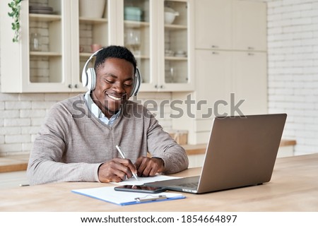 Happy african business man, black male student wearing headphones elearning on laptop computer sitting at kitchen table working from home office, learning online, studying remote training course. Royalty-Free Stock Photo #1854664897