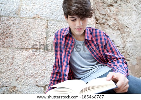 Close up of an attractive teenager student boy sitting on a college campus reading an open text book from school against an old stone building wall. Student outdoors lifestyle.