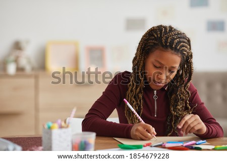 Front view portrait of teenage African-American girl doing homework while sitting at desk in home interior, copy space
