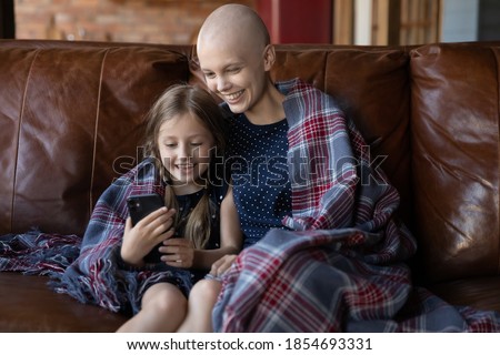 Look, mommy! Adorable little school age girl sitting on sofa under large shepherd plaid cuddling with hairless mother sick with cancer showing funny photo on cellphone screen making videocall together