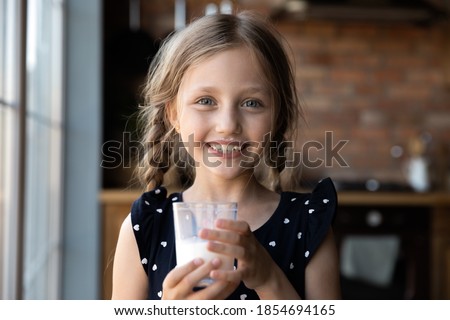 Tastes so good! Portrait of happy cheerful little girl holding glass of yoghurt looking at camera with milk mustache on face, cute child promotes healthy habit drink fresh farm dairy product every day